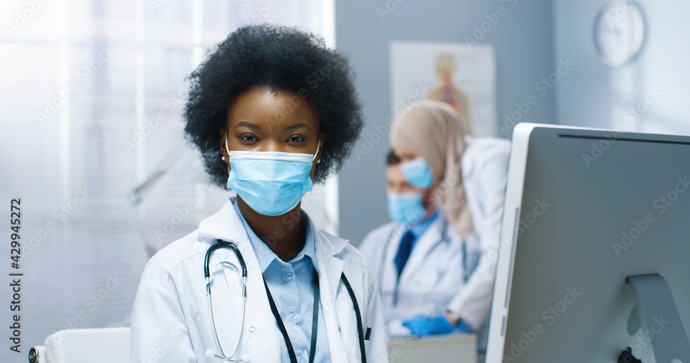 Close up portrait of cheerful young pretty African American woman in medical mask working on computer looking at monitor screen sitting in cabinet in hospital. Covid-19 pandemic, healthcare Close up portrait of cheerful young pretty African American woman in medical mask working on computer looking at monitor screen sitting in cabinet in hospital. Covid-19 pandemic, healthcare
