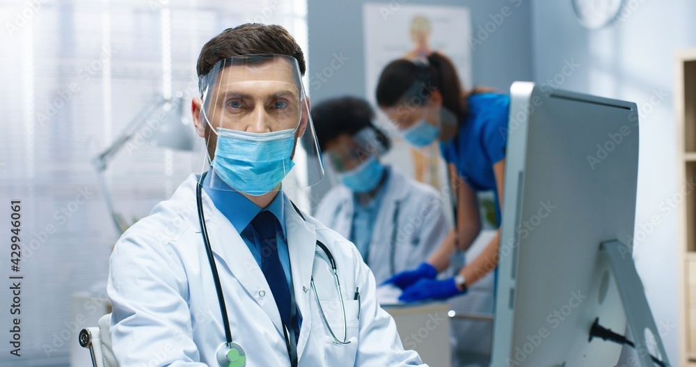 Close up portrait of handsome serious Caucasian male healthcare worker in mask and face shield working in hospital lab during coronavirus browsing on computer and looking at camera, clinic concept Close up portrait of handsome serious Caucasian male healthcare worker in mask and face shield working in hospital lab during coronavirus browsing on computer and looking at camera, clinic concept