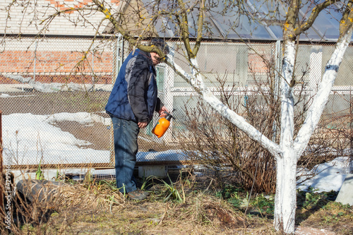 An elderly man is treating bushes and trees in the garden with a fungicidal