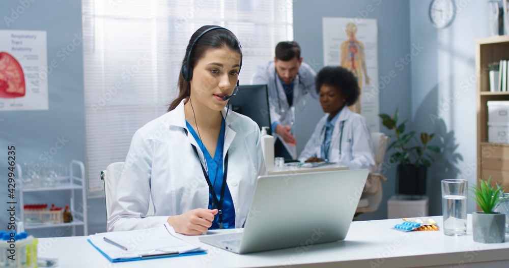 Caucasian young pretty female doctor having online video call consultation on laptop speaking in headset with patient sitting at table in hospital cabinet. Healthcare, Portrait, medical center concept Caucasian young pretty female doctor having online video call consultation on laptop speaking in headset with patient sitting at table in hospital cabinet. Healthcare, Portrait, medical center concept