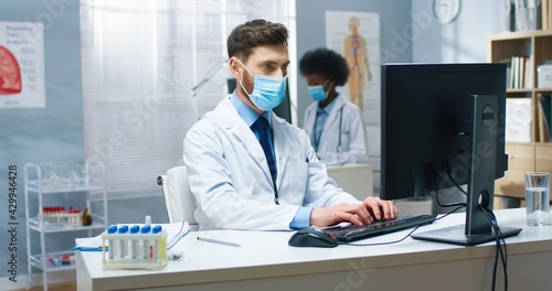 Wallpaper Mural Handsome young Caucasian experienced male doctor specialist in medical mask tapping on computer browsing online while sitting at workplace in hospital. Healthcare, medicine, medic concept, portrait Torontodigital.ca