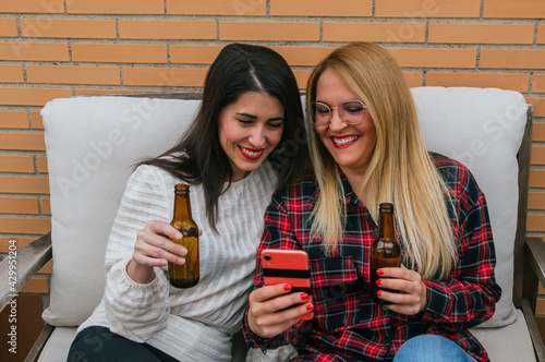 Two smiling young girls drinking beers and taking selfies with a mobile on a terrace