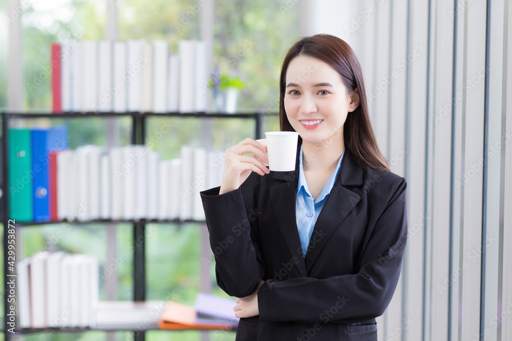 Asian business woman who wears blue shirt and black suit holds coffee cup in her hand and smile happily at work office as background. Asian business woman who wears blue shirt and black suit holds coffee cup in her hand and smile happily at work office as background.