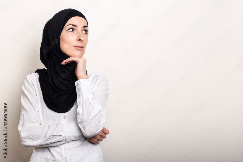 Young Muslim woman dressed in a white shirt and hijab holds her hand to her chin and looks to the side on a light background. Banner Young Muslim woman dressed in a white shirt and hijab holds her hand to her chin and looks to the side on a light background. Banner