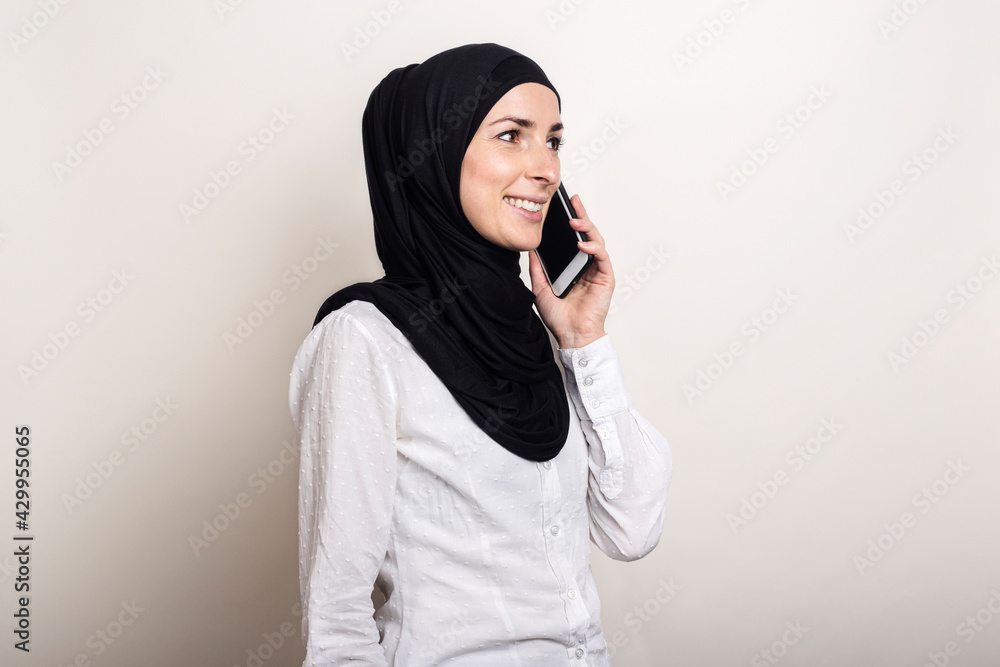 Muslim young woman in hijab talking on the phone and looking to the side on a light background. Banner Muslim young woman in hijab talking on the phone and looking to the side on a light background. Banner