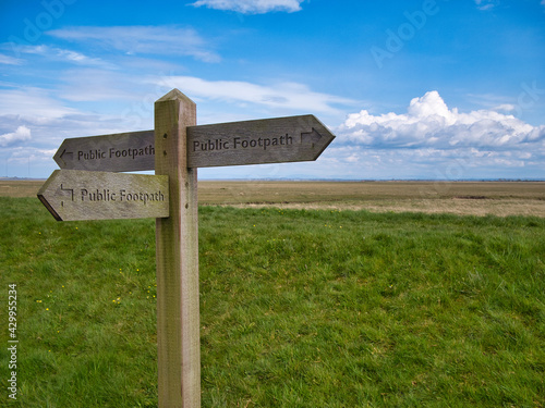 A wooden sign showing the directions of three public footpaths. Taken in a coastal setting on a sunny day with blue skies and white clouds.