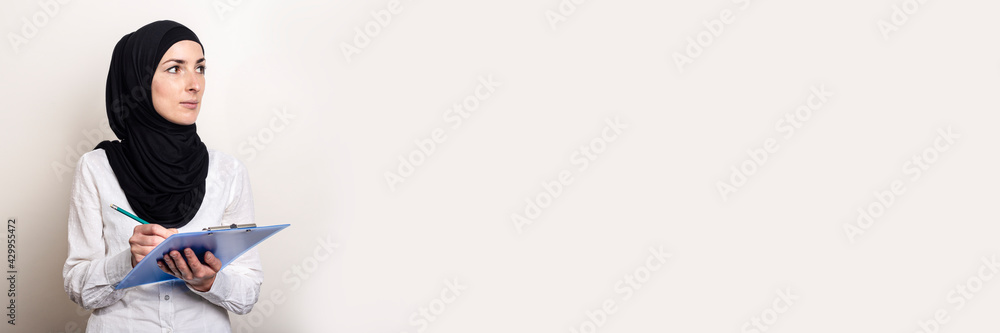 Young Muslim woman in a white shirt and hijab holds a clipboard and looks to the side. Auditor hiring. Banner Young Muslim woman in a white shirt and hijab holds a clipboard and looks to the side. Auditor hiring. Banner