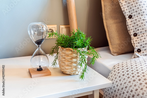 Bedside table with greenplant and hourglass on it. Interior details. light colours.