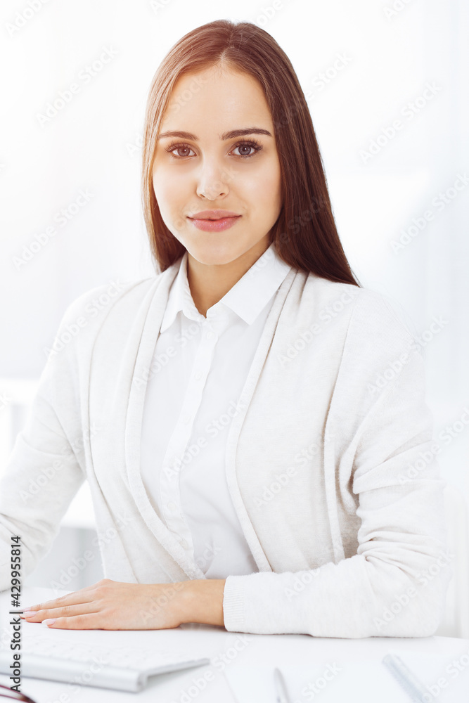 Young cheerful woman sitting at the desk with computer and looking at camera in sunny office. Headshot Young cheerful woman sitting at the desk with computer and looking at camera in sunny office. Headshot