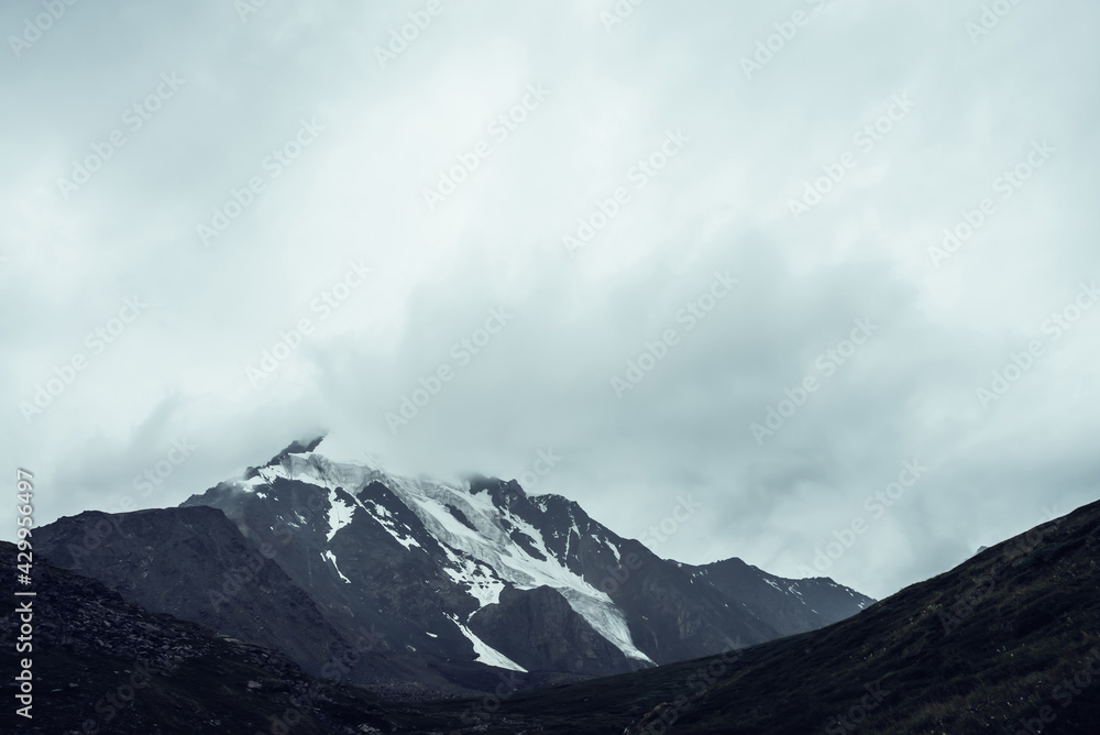 Minimalist monochrome atmospheric mountains landscape with big snowy mountain top in low clouds. Awesome minimal scenery with glacier on rocks. Black white high mountain pinnacle with snow in clouds.
