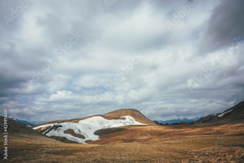 Fototapeta Naklejka Na Ścianę i Meble -  Sunny mountain desert relief with glacier in sunlight under low cloudy sky. Scenic highlands landscape with small glacier high in mountains on hills. Minimalist mountain scenery of highland desert.