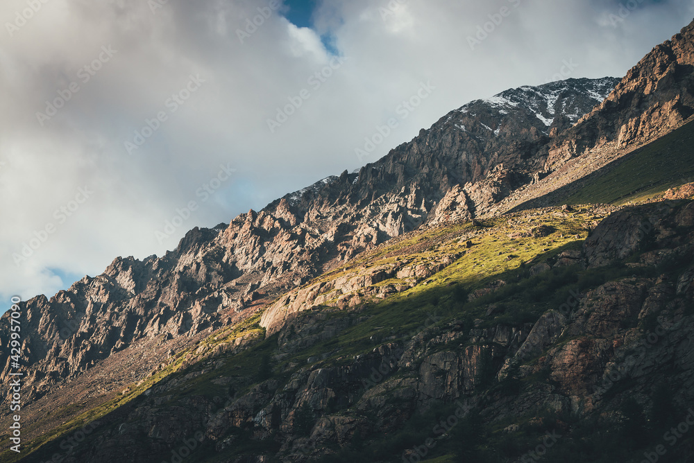 Beautiful mountain landscape with high green mountainside with sharp rocks in sunlight. Atmospheric mountain scenery with steep slope in sunshine. Awesome sunny view to great mountain and low clouds.