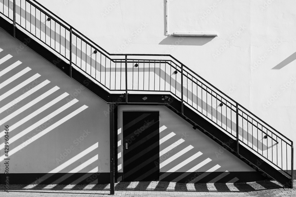 Fototapeta premium Eine Außentreppen zieht sch diagonal an der Hauswand entlang und wirft Schatten auf die Wand und den gepflasterten Boden - in schwarzweiß.