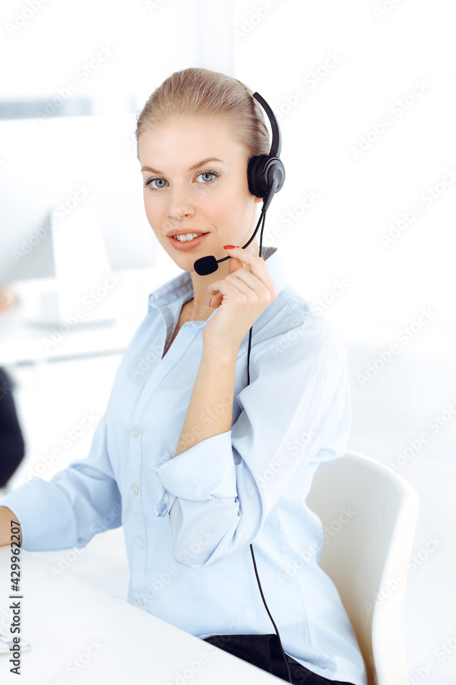 Blond woman call operator is using computer and headset for consulting clients online. Group of diverse people working as customer service occupation. Business concept Blond woman call operator is using computer and headset for consulting clients online. Group of diverse people working as customer service occupation. Business concept