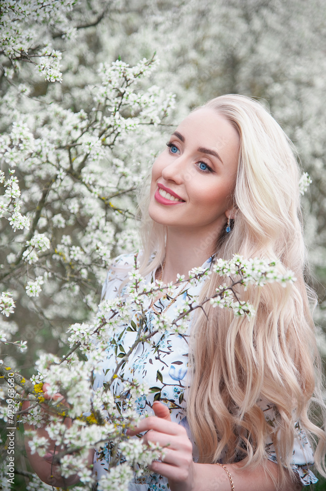 .A beautiful blonde girl with long hair, bright blue eyes, with clean skin and natural make-up against the background of densely blooming white flowers of spring trees. Outdoors.