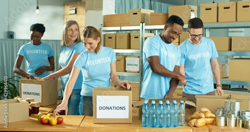 Mixed-race happy positive male and female different volunteers working in charity organization center preparing and packing social help box, donations box, volunteering and donating concept