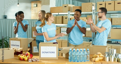 Joyful happy diverse Caucasian and African American men and women volunteers standing in social organization warehouse clapping hands and giving high five. Donations, charity work, volunteering