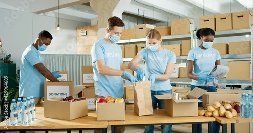 Group of Caucasian and African American male and female volunteers putting food in packet in shelter as charity workers and members of community work giving food to poor during coronavirus pandemic