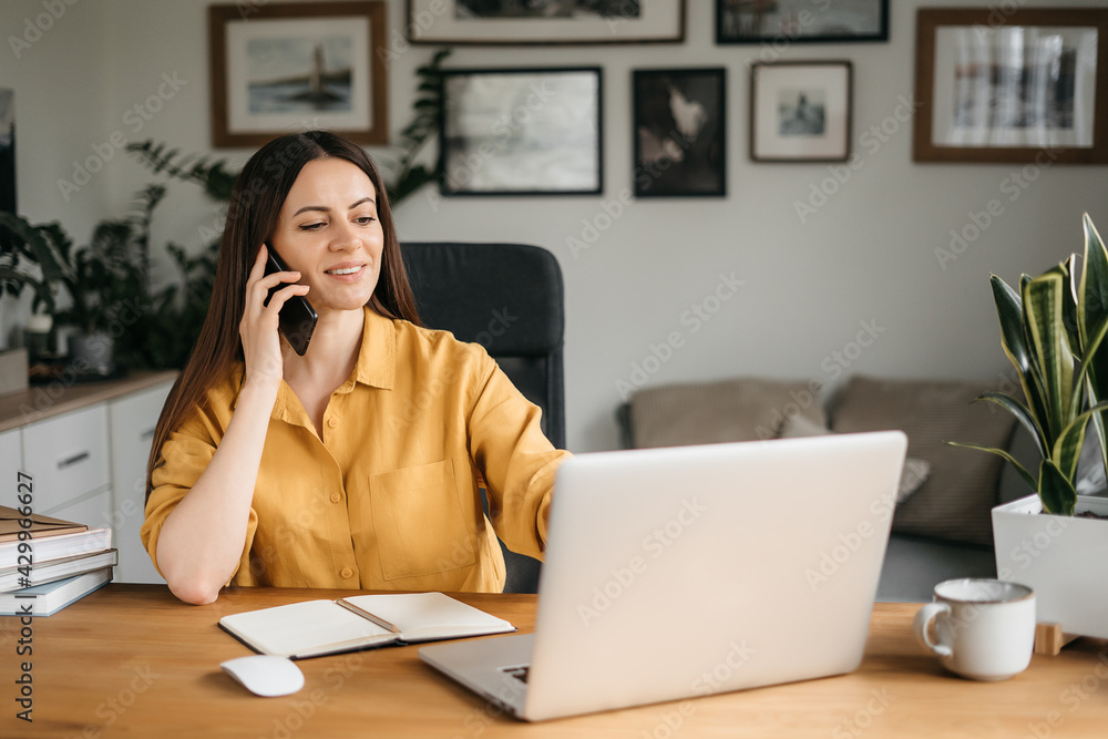 Smiling business woman talking on a mobile phone and using a laptop while working at home, looking at the screen, reading a message, a young female employee advises a client, telephone conversations Smiling business woman talking on a mobile phone and using a laptop while working at home, looking at the screen, reading a message, a young female employee advises a client, telephone conversations