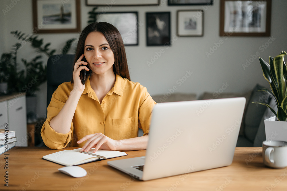 Smiling business woman talking on a mobile phone and using a laptop while working at home, looking at the screen, reading a message, a young female employee advises a client, telephone conversations Smiling business woman talking on a mobile phone and using a laptop while working at home, looking at the screen, reading a message, a young female employee advises a client, telephone conversations