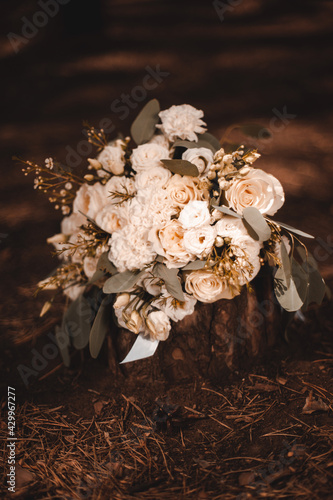 A bouquet of chic white and beige roses tied with a ribbon lies on a stump in the sun at sunset.