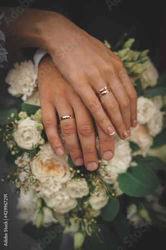 The hands of the newlyweds with gold wedding rings. One on top of the other is a cross on a bouquet of white wedding flowers.