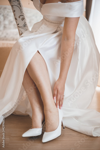 A beautiful young bride in a white long dress touches her delicate feet in white shoes with beautiful hands.