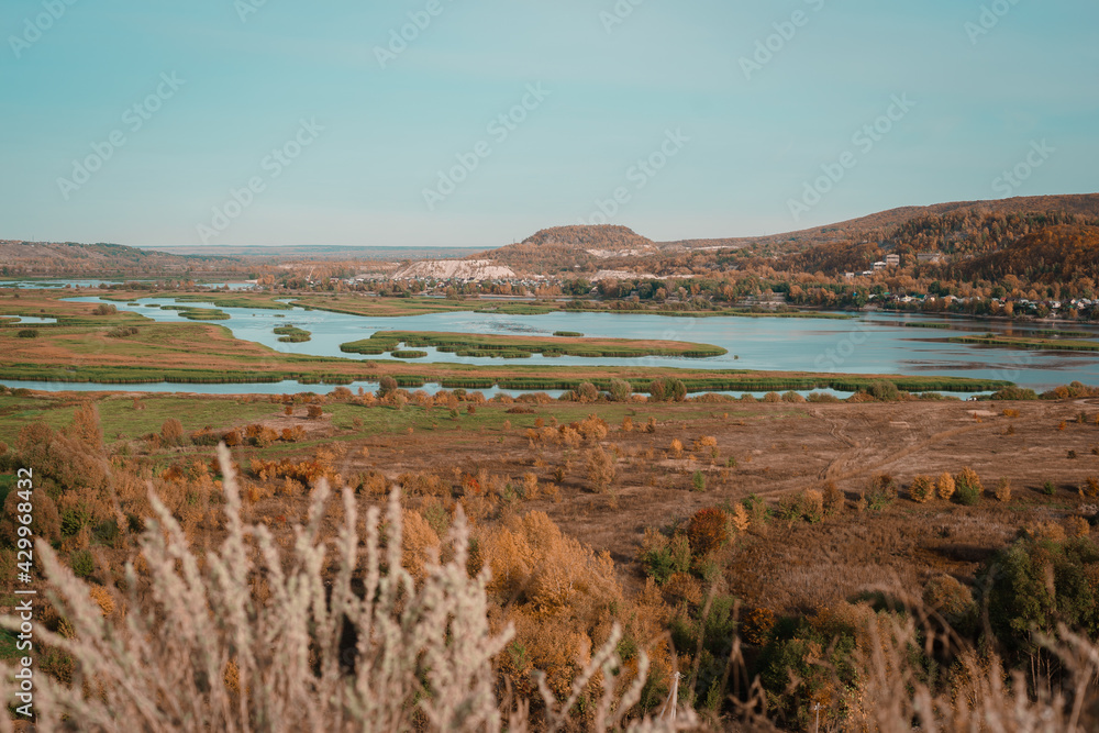 Beautiful autumn landscape. River valley. On the right is a large mound. Hill. A winding river. The colors of autumn. Brown, Red, Orange, Yellow, Turquoise. Blue lakes. 