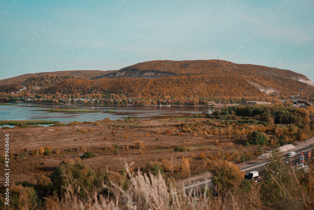 Beautiful autumn landscape. River valley. On the right is a large mound. Hill. A winding river. The colors of autumn. Brown, Red, Orange, Yellow, Turquoise. Blue lakes. A winding highway.