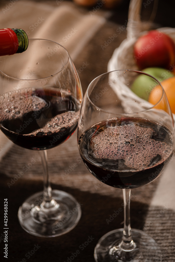 Red wine in large glass glasses foams on a brown tattered blanket. Next to it are ripe red, orange and green fruits in a basket. Red wine in large glass glasses foams on a brown tattered blanket. Next to it are ripe red, orange and green fruits in a basket.