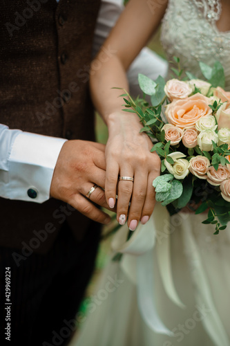 The hands of the newlyweds with gold wedding rings. One on top of the other is a cross on a bouquet of wedding flowers.
