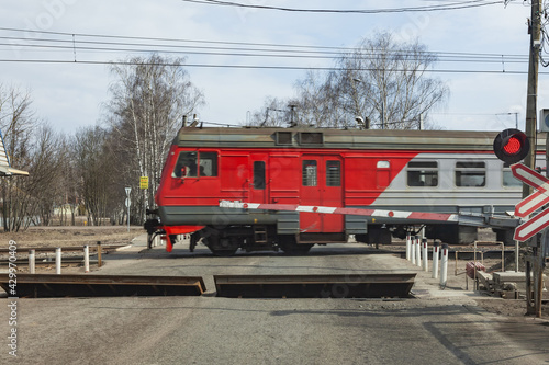 There are cars at the railway crossing. The barrier is lowered, the red signal of the semaphore is lit. A commuter train passes by. Russia, Moscow April 2021