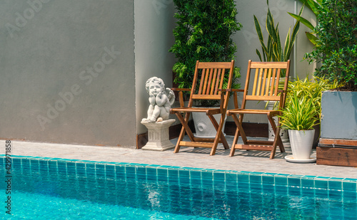 Two vintage wooden chairs together beside swimming pool with small garden decoration by many tree in pots, daylight image.