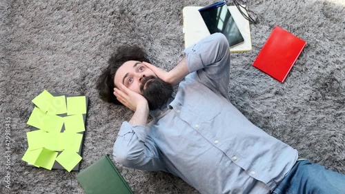 Young bearded man lying on the floor, surrounded by computer and documents, the man is very tired and exhausted, laptop is full of reminder notes.