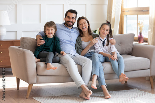 Portrait of happy young Caucasian family renters with two little children relax on couch in new modern home. Smiling parents with small kids sit rest on sofa in renovated design house. Rent concept.