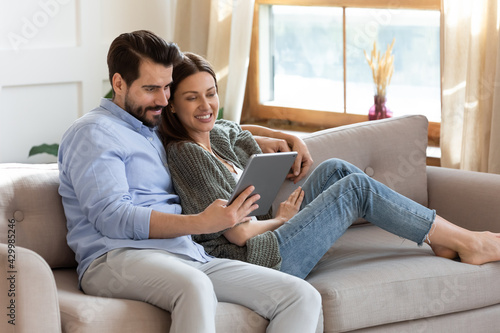Happy young Caucasian couple renters relax on sofa at home use modern tablet gadget browsing internet. Smiling millennial man and woman look at pad screen talk speak on video webcam call on gadget.