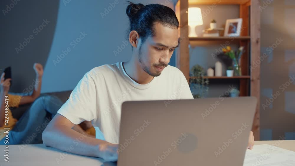 Freelance asian couple man and woman in casual husband focus work in laptop computer at night wife take a rest feel relax wear headphones play music on smartphone lay down and dance on couch at home. Freelance asian couple man and woman in casual husband focus work in laptop computer at night wife take a rest feel relax wear headphones play music on smartphone lay down and dance on couch at home.