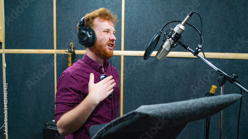 Expressive bearded man with curly ginger hair in headphones at recording studio stay opposite a microphone and performs like voice actor