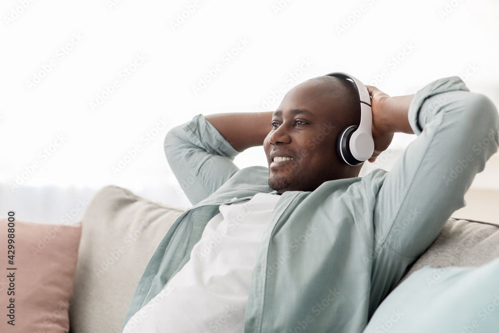 Fototapeta premium African american middle-aged man in wireless headphones listening to music on sofa in living room, copy space