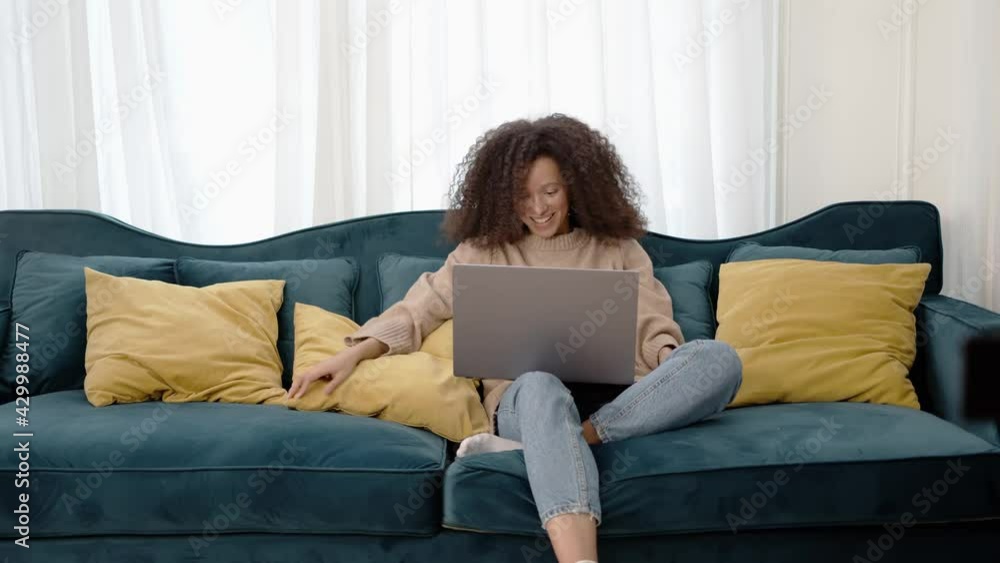 Young emotional american black woman, cheerful hands waving, video call on the laptop. Gesturing and smiling. Curly afro hairstyle and long sofa with pillows in living room Young emotional american black woman, cheerful hands waving, video call on the laptop. Gesturing and smiling. Curly afro hairstyle and long sofa with pillows in living room