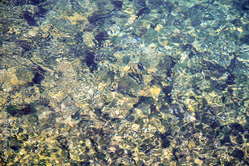 The pebbles at the bottom of the stream shine through and shimmer with light at the bottom of the stream with crystal clear water. Texture. Transparent. Background. Abstract