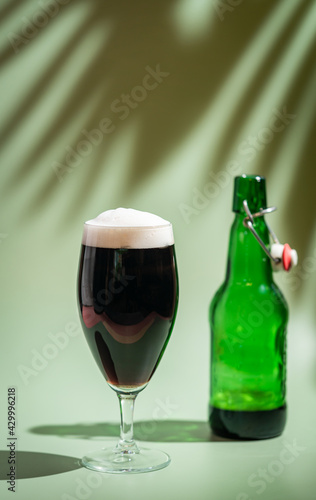 Glass of dark beer with foam head on colorful green background, bright shadows of palm leaves