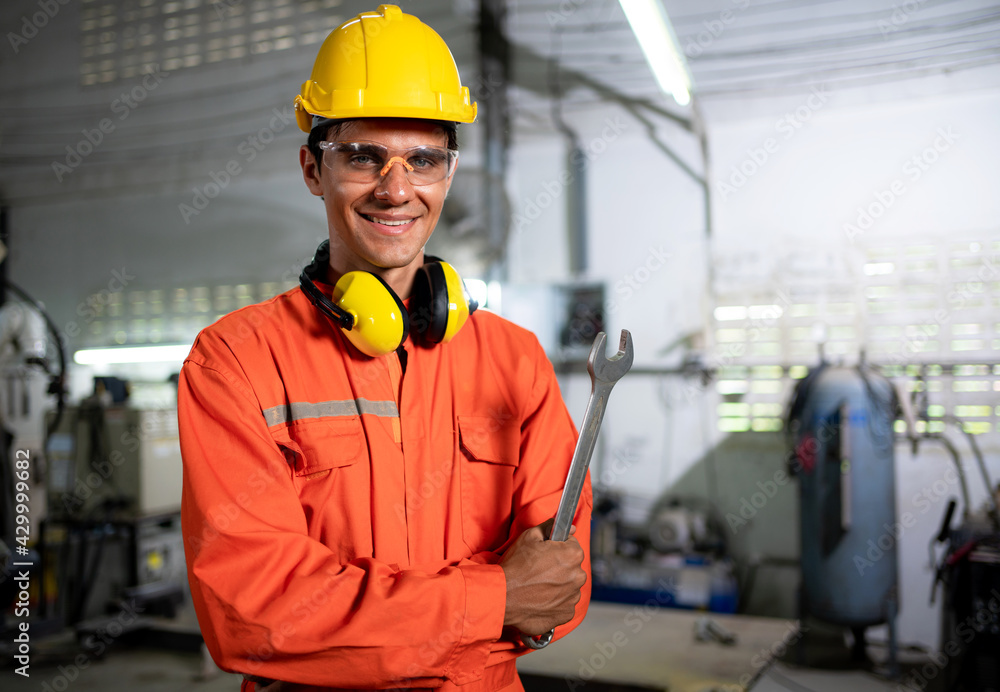 Portrait of young engineer standing and work in the robot factory. Wearing orange uniform, safety helmet crossed arms and smile with copy space. Holding wrench. Technology and lifestyle concept Portrait of young engineer standing and work in the robot factory. Wearing orange uniform, safety helmet crossed arms and smile with copy space. Holding wrench. Technology and lifestyle concept