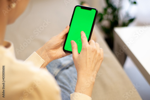 Woman at home, on the couch, using a smartphone with a green mockup screen. Girl using a mobile phone on the Internet. Point of view shot