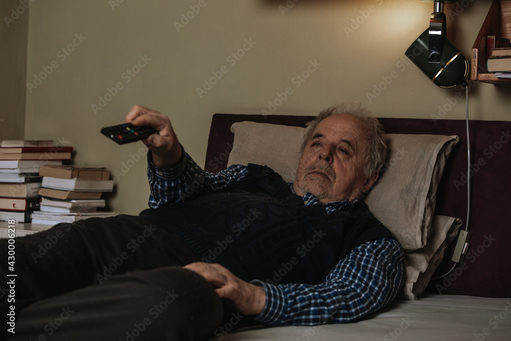 Senior man napping while lying down in bed and watching TV with a television remote, resting in a bed