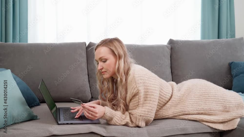 Beautiful blonde girl makes online payments with a bank card, using a modern laptop at home, lying on the couch and smiling. Finance, online shopping. Beautiful blonde girl makes online payments with a bank card, using a modern laptop at home, lying on the couch and smiling. Finance, online shopping.