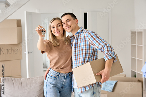 Happy young couple home owners holding keys in new home. Smiling independent millennial man and woman first time homeowners carrying boxes on moving day. Mortgage loan, new house ownership concept.