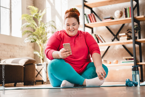Relaxing fat plus size caucasian woman resting after hard fitness training at home using smart phone. Chubby obese woman wants to lose weight and burn calories