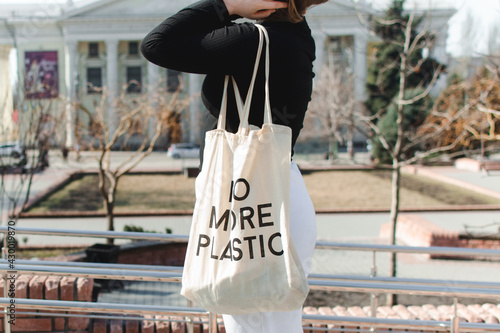 Young girl in monochrome clothes with cotton cloth grocery bag with inscription no more plastic stands against background of city street. Copy space for text. Eco friendly zero waste natural concept