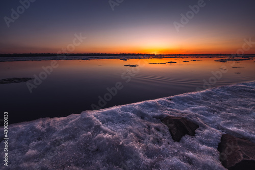 A beautiful sunset over a large river with floating ice and rocks on the shore. Spring season.